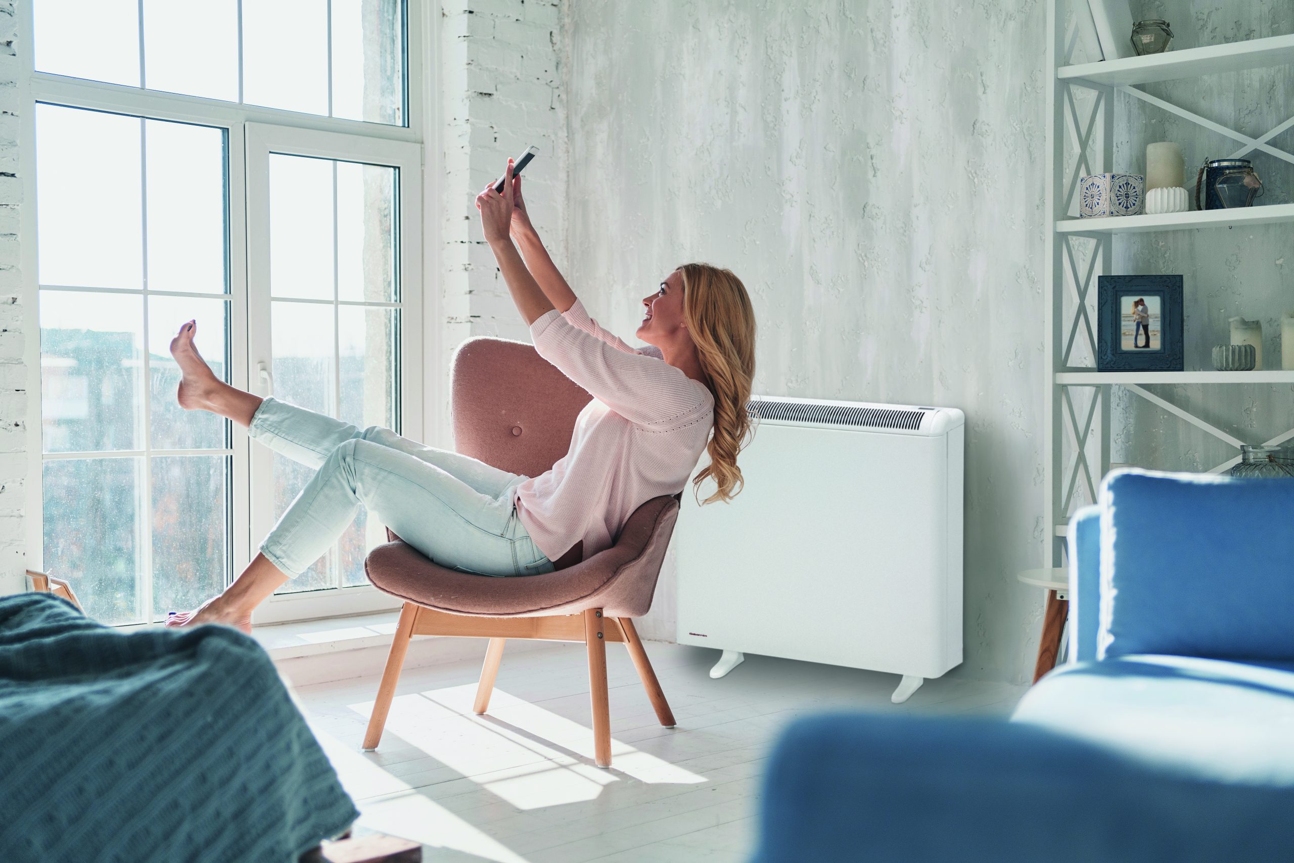 Happy woman on phone while sitting on wooden chair in living room, with Elnur's PROSSH Storage Heater in background